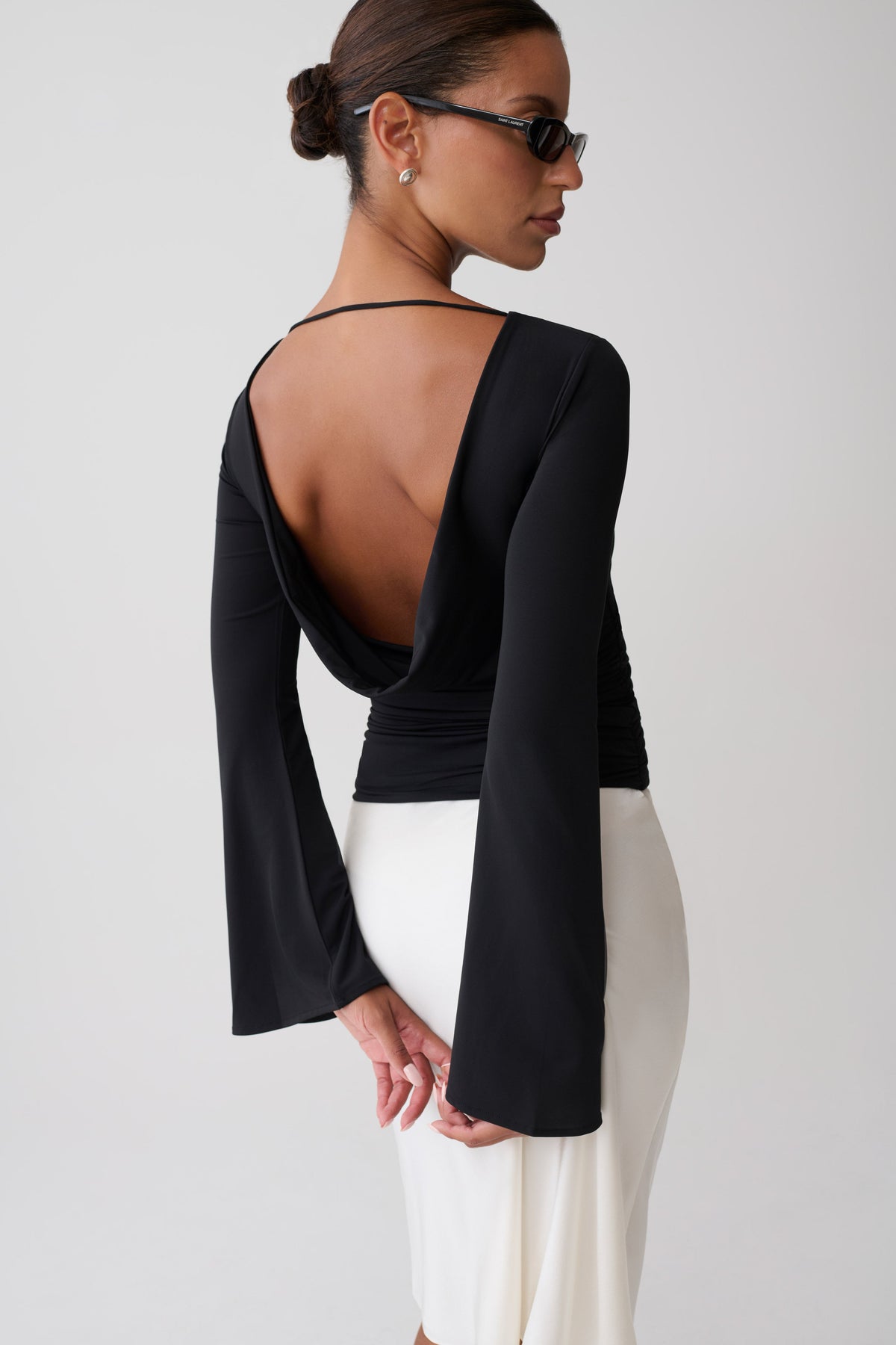 A woman shown from the back wearing a black long-sleeve top with a dramatic open back and white skirt against a light gray studio background.