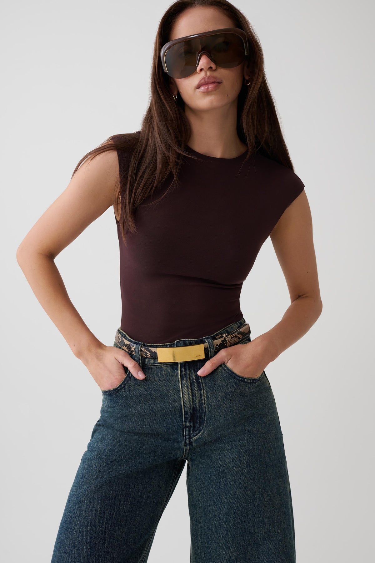 A woman modeling a fitted sleeveless dark brown bodysuit with blue jeans and a gold belt, wearing oversized dark sunglasses against a light gray studio background.