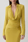 A woman wearing a fitted long-sleeve dress in a rich mustard yellow. The dress features a draped cowl neckline, a cinched waist, and a deep V-neckline.