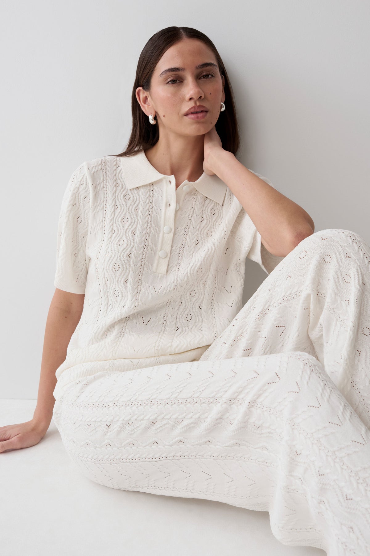 A woman wearing a matching cream-colored knit two-piece set consisting of a short-sleeve polo shirt and wide-leg trousers, seated against a plain light gray wall.