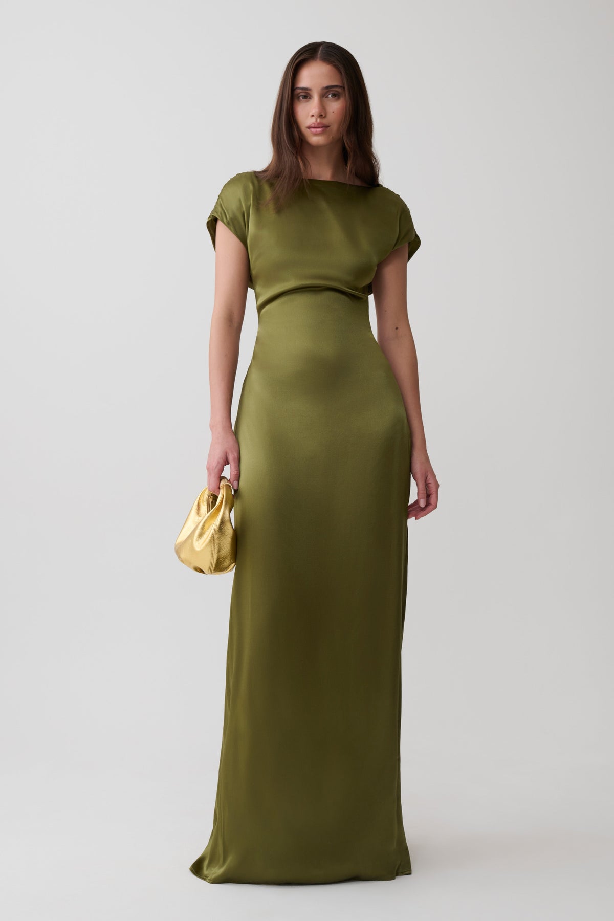 A woman modeling a floor-length olive green satin dress with short cap sleeves and a draped waist detail, holding a small metallic gold clutch against a light gray studio background.