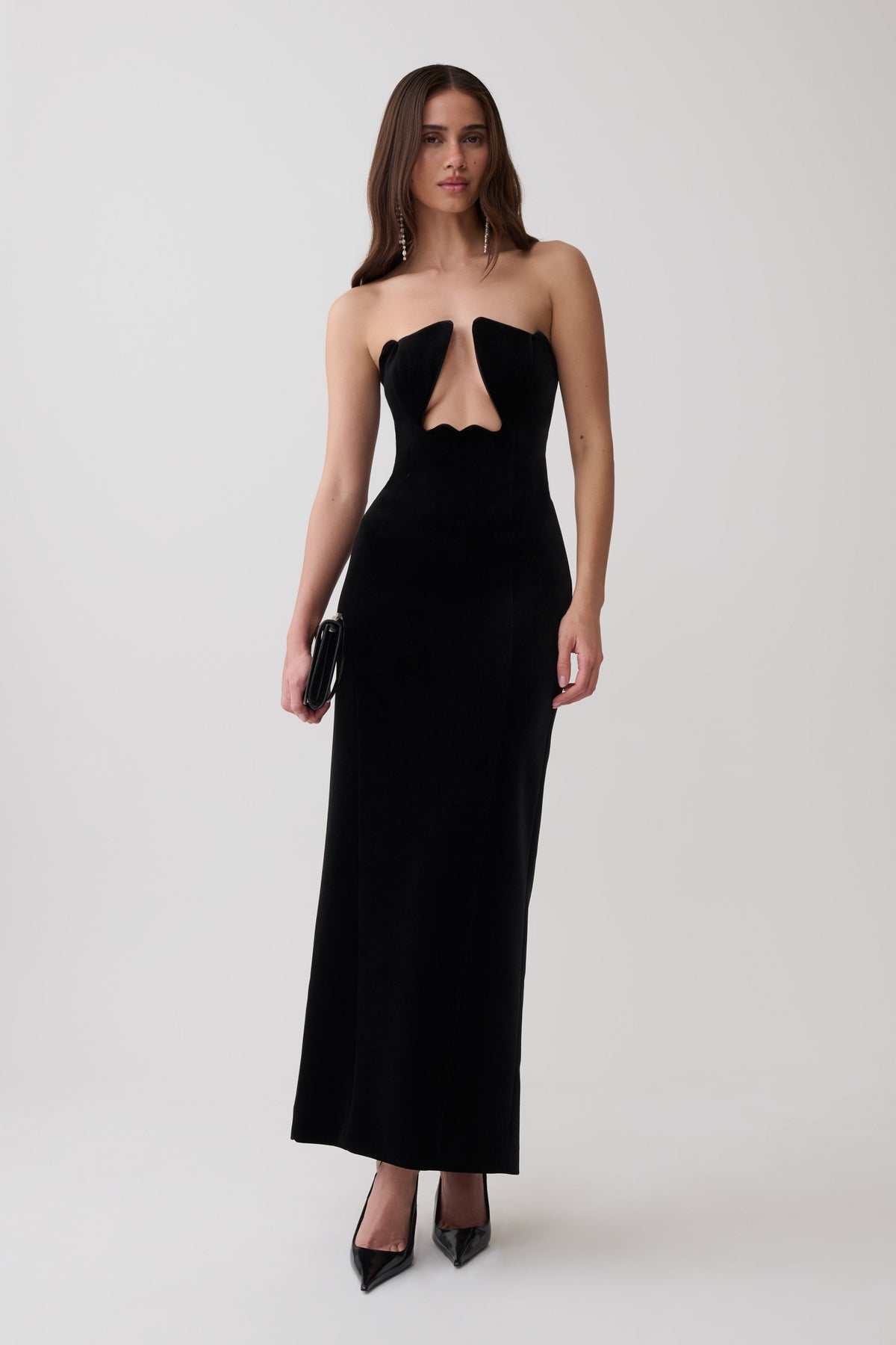 A woman modeling a strapless black velvet maxi dress with a sweetheart neckline and a keyhole cutout at the waist, paired with black pointed-toe heels against a light gray studio background.
