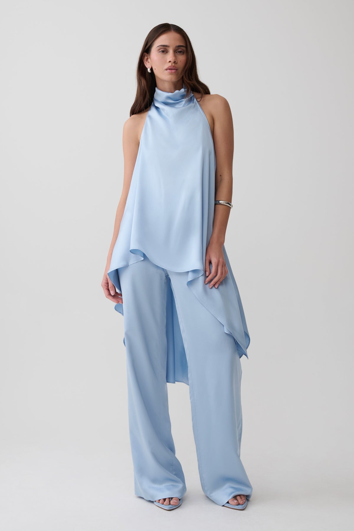 A model wearing a light blue sleeveless draped top and matching wide-leg pants, photographed against a plain light gray studio background.