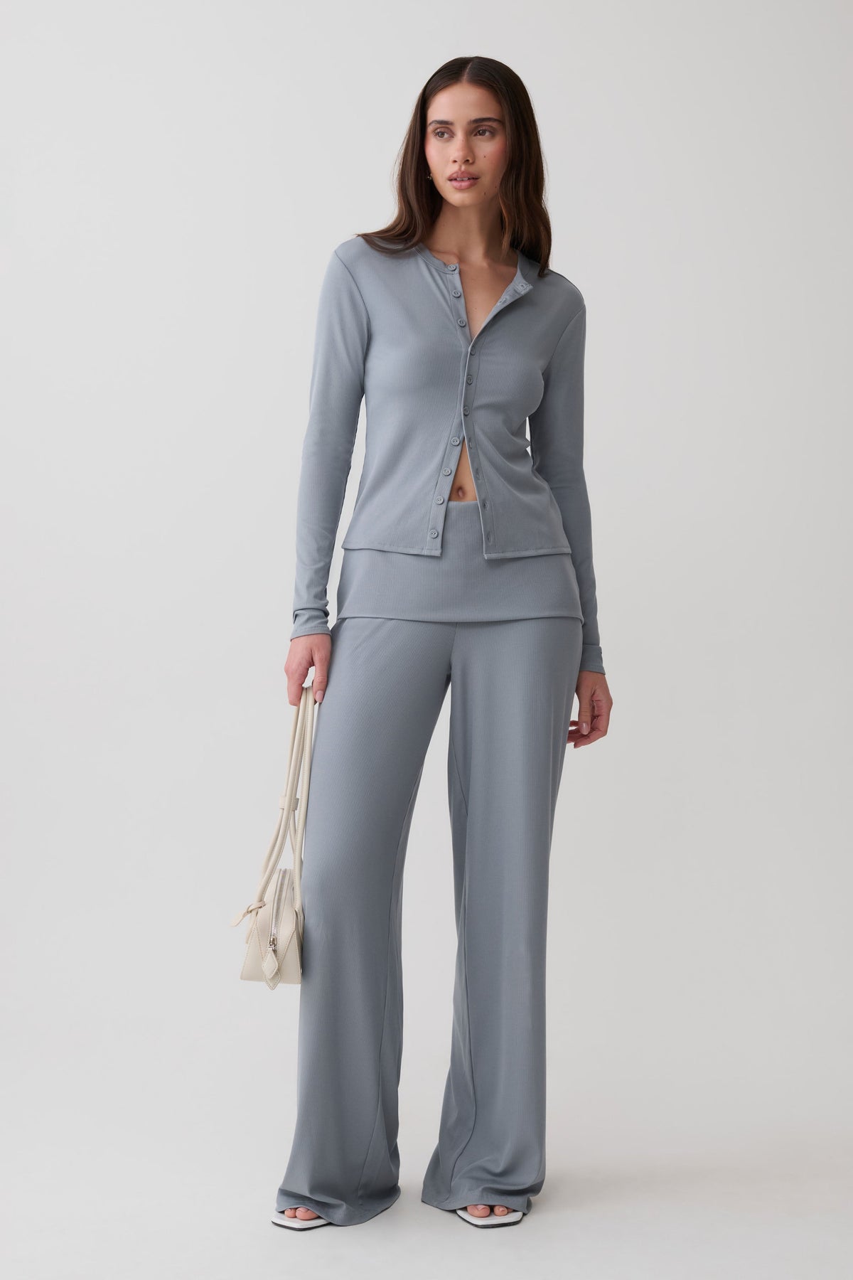 A woman modeling a matching two-piece loungewear set in a dusty slate gray color, consisting of a long-sleeve button-front top and wide-leg pants, holding a small cream-colored shoulder bag against a light gray studio background.